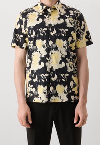 Camisa de manga corta con botones, presenta un fondo negro con grandes patrones florales amarillos, hecha de una tela suave con un cuello estructurado.