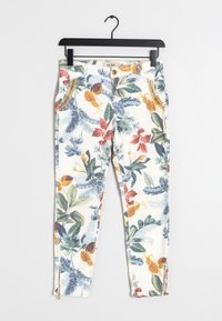 Pantalones con estampado floral en blanco, con hojas y insectos coloridos. Incluyen cremalleras laterales en los tobillos y un cierre de botón dorado.