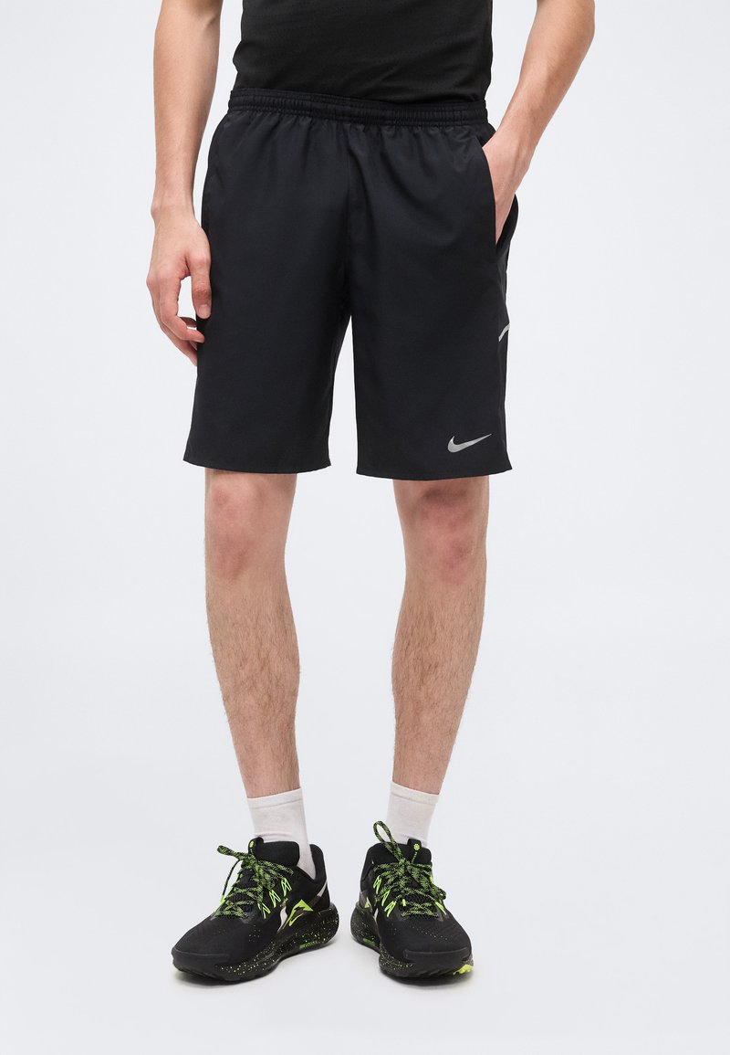 Άνδρας που φοράει μαύρα σορτς Nike, λευκές κάλτσες και μαύρα αθλητικά παπούτσια με νέον πράσινες λεπτομέρειες, στέκεται με το ένα χέρι στην τσέπη.