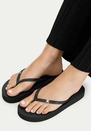 Piedi con infradito platform neri con logo argento sulle fasce, abbinati a pantaloni neri plissettati su sfondo uniforme.