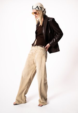 Vrouw die beige wijde broek, bruin topje, donkerleren jas, beige hoofddoek met tekst, zonnebril en open-toe hakken draagt, staand met handen in de zakken.