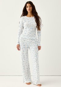 Γυναίκα όρθια ξυπόλητη, φοράει λευκό ριμπ μακρυμάνικο τοπ και παντελόνι με μικρό μπλε ανθισμένο μοτίβο μπροστά σε ένα απλό φόντο.