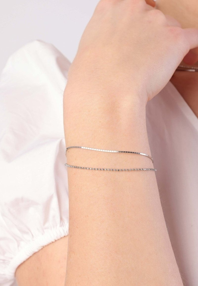 Silver bracelet with two delicate chains; one features small round beads and the other a flat, textured surface. Worn on a wrist.