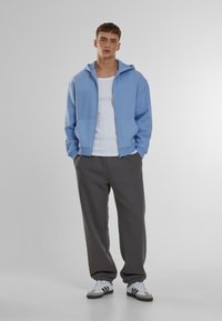Sudadera de cremallera azul claro sobre una camiseta blanca sin mangas, combinada con pantalones de chándal gris oscuro y zapatillas blancas con rayas negras. Textura de tela suave.