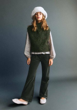 Gilet en tricot vert sur une blouse imprimée, associé à un pantalon sombre et des chaussons en fausse fourrure clairs. Un chapeau en fourrure blanche complète le look.