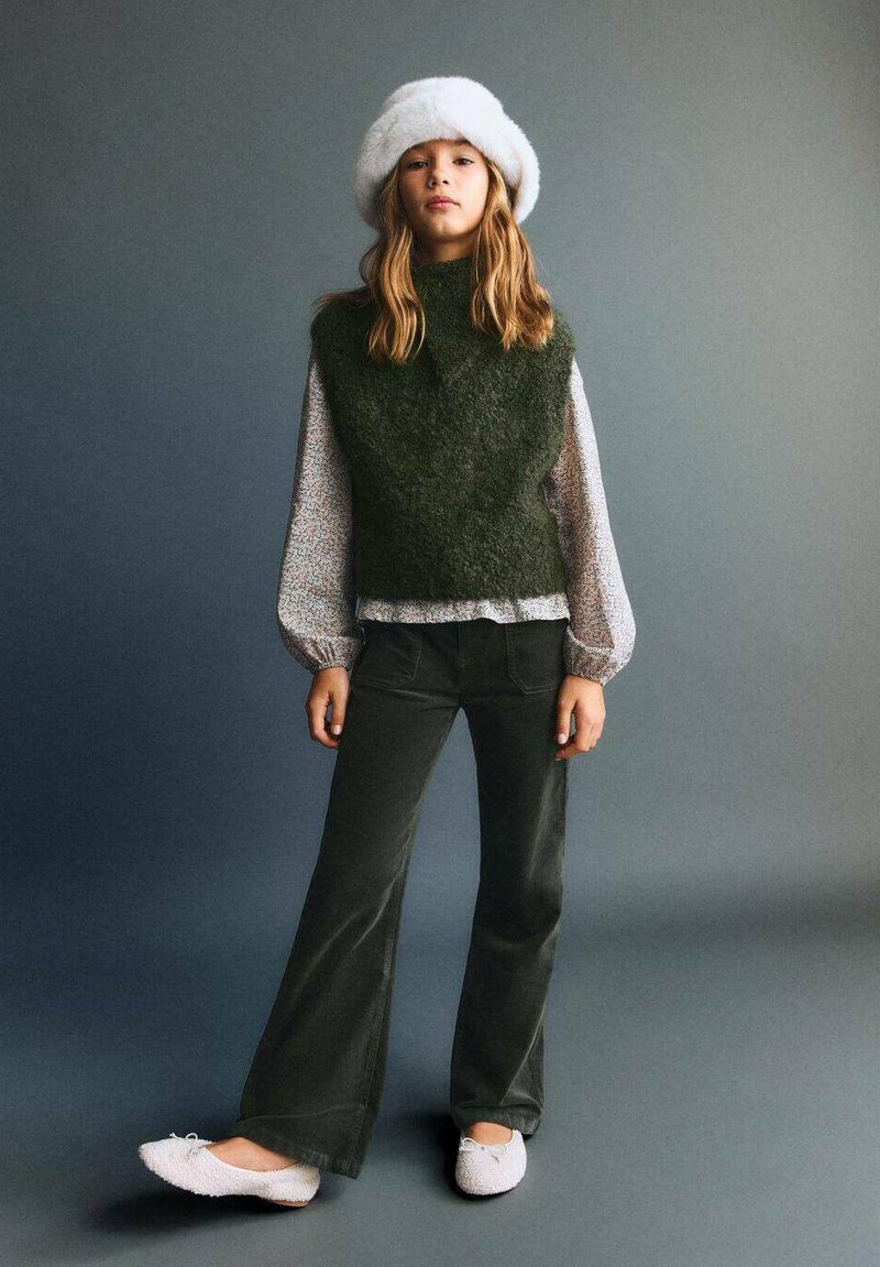 Grüner gestrickter Weste über einer gemusterten Bluse, kombiniert mit dunklen Hosen und leichten, flauschigen Hausschuhen. Ein weißer Pelzhut rundet den Look ab.