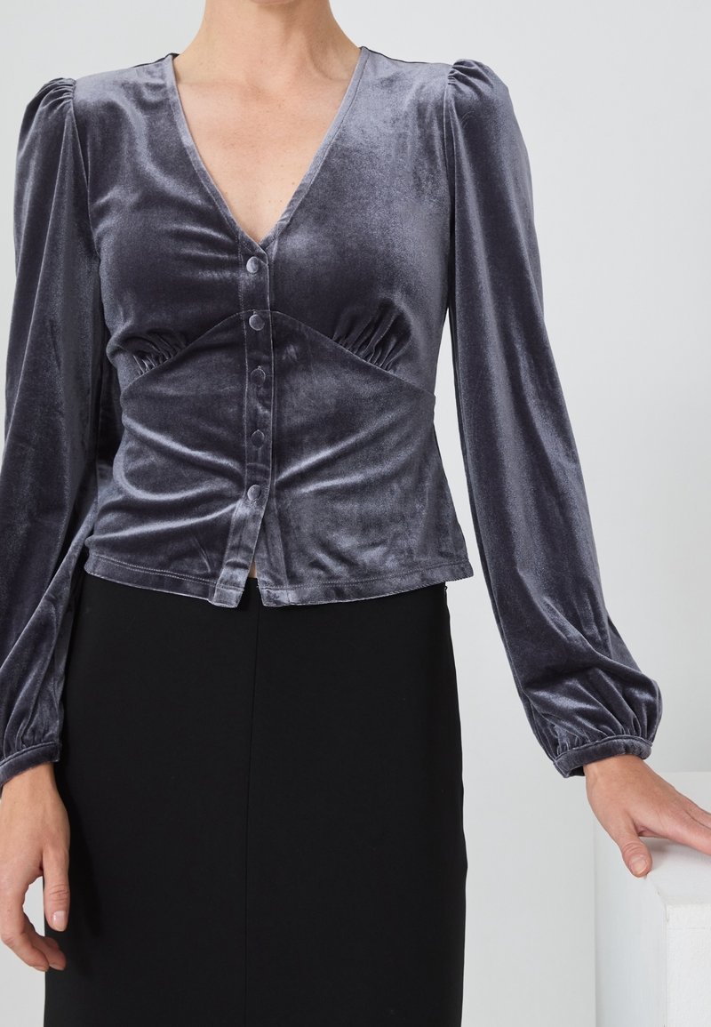 Femme portant un haut boutonné en velours gris foncé avec des manches longues bouffantes et une jupe noire, debout avec une main posée sur une surface blanche.