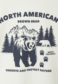 Graphic t-shirt featuring a brown bear design, navy print on cream fabric, with mountains, trees, and text: “North American Brown Bear.”