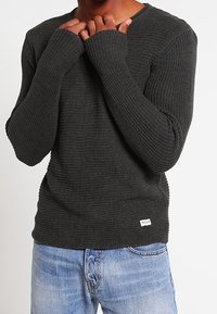 Dunkelgrauer Strickpullover mit langen Ärmeln und Daumenlöchern, der ein strukturiertes Muster aufweist. Kombiniert mit hellblauen Jeans.