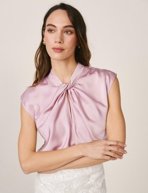 Mujer con cabello largo y castaño que lleva una blusa sin mangas de color rosa claro brillante con un detalle de nudo retorcido en el escote y una falda blanca con textura.