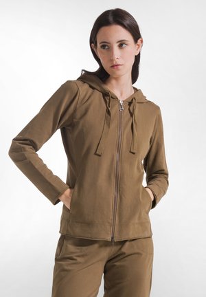 Donna che indossa una felpa marrone con zip e pantaloni abbinati, in piedi con le mani nelle tasche, guardando leggermente di lato.