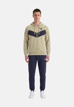 Giovane uomo che indossa una felpa beige con zip con motivo a chevron scuro, pantaloni da jogging blu navy e scarpe da ginnastica bianche, in piedi davanti a uno sfondo semplice.