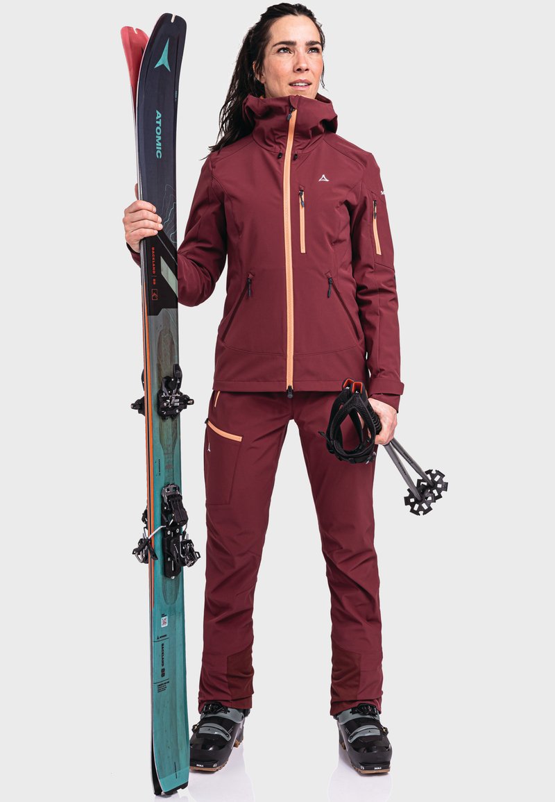 Bordeauxfarbene Skijacke mit hellorangefarbenen Akzenten, kombiniert mit passender Hose. Skier und schwarze Skistöcke in den Händen gehalten. Wasserdichtes Material.