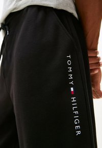 Pantalones de chándal negros hechos de una tela suave, con un corte holgado y bolsillos laterales. Logo blanco de "TOMMY HILFIGER" con acentos de color en la pierna.