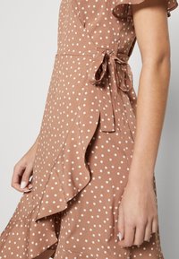 Vestido marrón con lunares que presenta un diseño envolvente, un dobladillo con volantes y un lazo lateral. Confeccionado en una suave tela, su textura sin costuras mejora la comodidad.