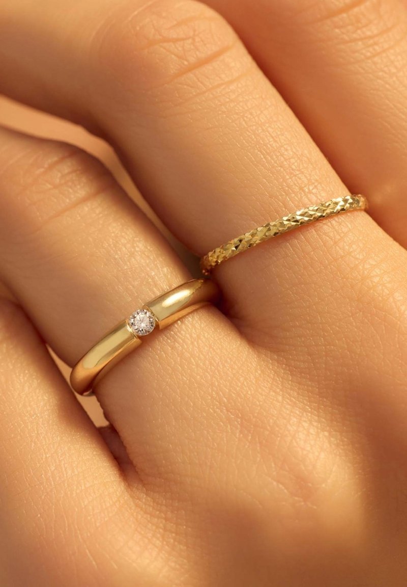 Goldringe an einer Hand. Einer ist glatt mit einem zentralen Diamanten, der andere hat ein strukturiertes, geflochtenes Muster. Hautfarbe ist darunter sichtbar.
