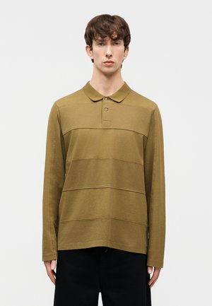 Giovane uomo con capelli castani corti che indossa una polo verde oliva a maniche lunghe e pantaloni neri, in piedi davanti a uno sfondo chiaro e semplice.