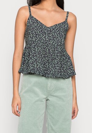 Woman wearing a black camisole with green floral print and light green corduroy pants against a plain background.