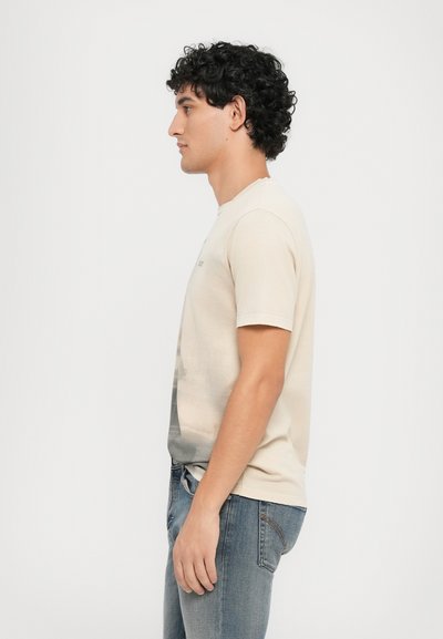 Beige bomulds t-shirt med et fotografisk grafisk print på fronten, rund halsudskæring og korte ærmer. Bæres med blå jeans.