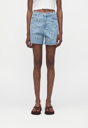 Λεπτό άτομο που φοράει ανοιχτό μπλε τζιν σορτς με μικρά σχέδια μαργαρίτας, λευκό κοντό τοπ και καφέ πλατφόρμες σανδάλια, στέκεται μπροστά σε λευκό φόντο.