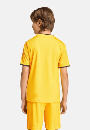 Enfant portant un maillot de sport jaune uni et un short, debout de dos sur un fond blanc uni.