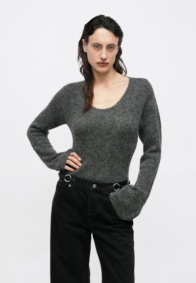 Szürke kötött pulóver mély kivágással és megnyújtott ujjal, magas derekú fekete farmerrel, amely kör alakú díszítésekkel van ellátva.