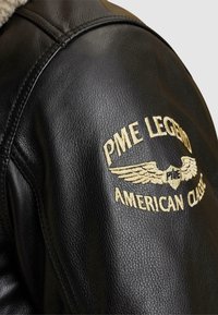 Črna usnjena jakna z šivanimi detajli. Vezeni logo na rami z obliko kril v zlati barvi, ki označuje ime blagovne znamke in stil.