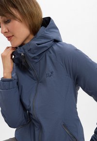 Veste zippée bleue avec un col haut, arborant un logo brodé, des poignets élastiques et une texture lisse et mate.