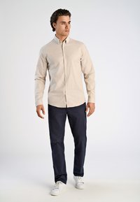 Helle beige Button-Up-Hemd, marineblaue Hose, weiße Sneakers. Das Hemd hat einen klassischen Kragen und lange Ärmel mit einer subtilen Textur.
