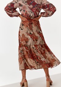 Langer, gestufter Rock aus einem gemusterten Stoff mit roten, beigen und blauen Blumenmotiven. Leicht durchsichtig mit einem Rüschensaum und bronzefarbenen Absätzen.