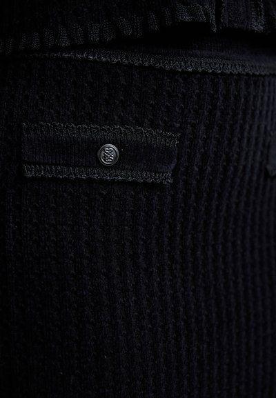 Un material texturat negru are o margine ondulată și un mic accent cu un buton negru pe o secțiune peticită, subliniind detaliul complex.
