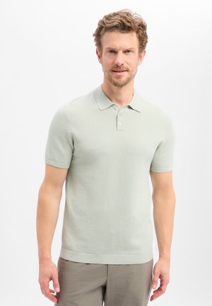 Man met lichtbruin krullend haar, gekleed in een getextureerd lichtgrijs kortemouws poloshirt en grijze broek, staand tegen een witte achtergrond.