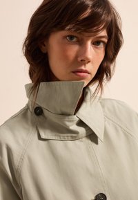 Lichtgroene trenchcoat met een hoge kraag, voorzien van knopen op de revers en een geweven stof, benadrukt door nauwkeurige stikseldetails.