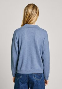 Femme aux longs cheveux blonds portant un pull bleu clair et un jean bleu foncé, vue de dos sur un fond uni.