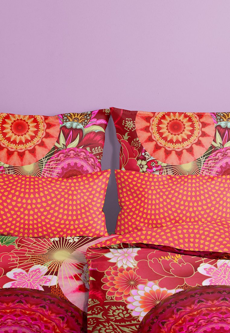 Helle, gemusterte Bettwäsche mit lebendigen roten und rosa Blumenmotiven, Herzmustern auf den Kissenbezügen, vor einem lila Wandhintergrund.