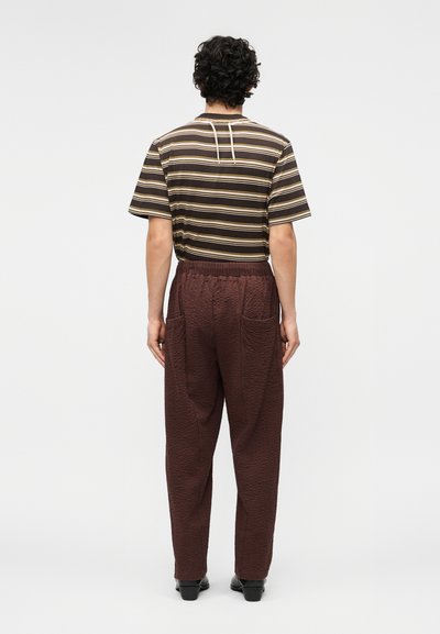 Άνδρας με σγουρά μαλλιά που φοράει ριγέ καφέ και μπεζ κοντομάνικο πουκάμισο και υφασμένα σκούρα καφέ παντελόνια, στέκεται γυρισμένος προς τα πίσω.