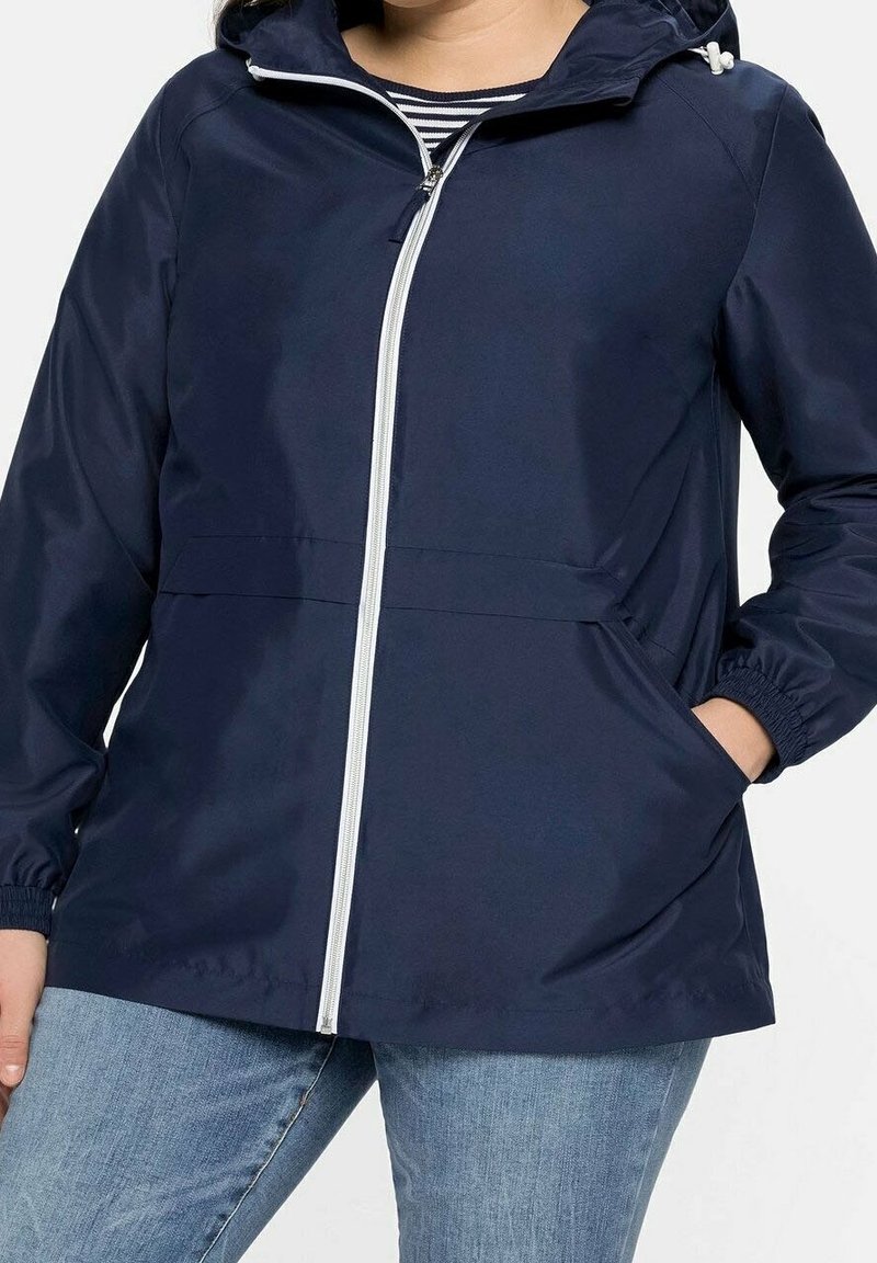 Giacca impermeabile leggera blu navy con cappuccio, dotata di una cerniera bianca, polsini elastici e tasche laterali. Texture liscia e design casual.