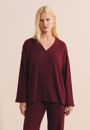 Donna che indossa una camicia ampia a costine, rosso scuro, abbottonata e pantaloni coordinati a costine, in piedi contro uno sfondo neutro.