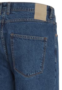Nærbillede af baglommen og linningen på blå denimjeans med brun syning og tannet læderlap.