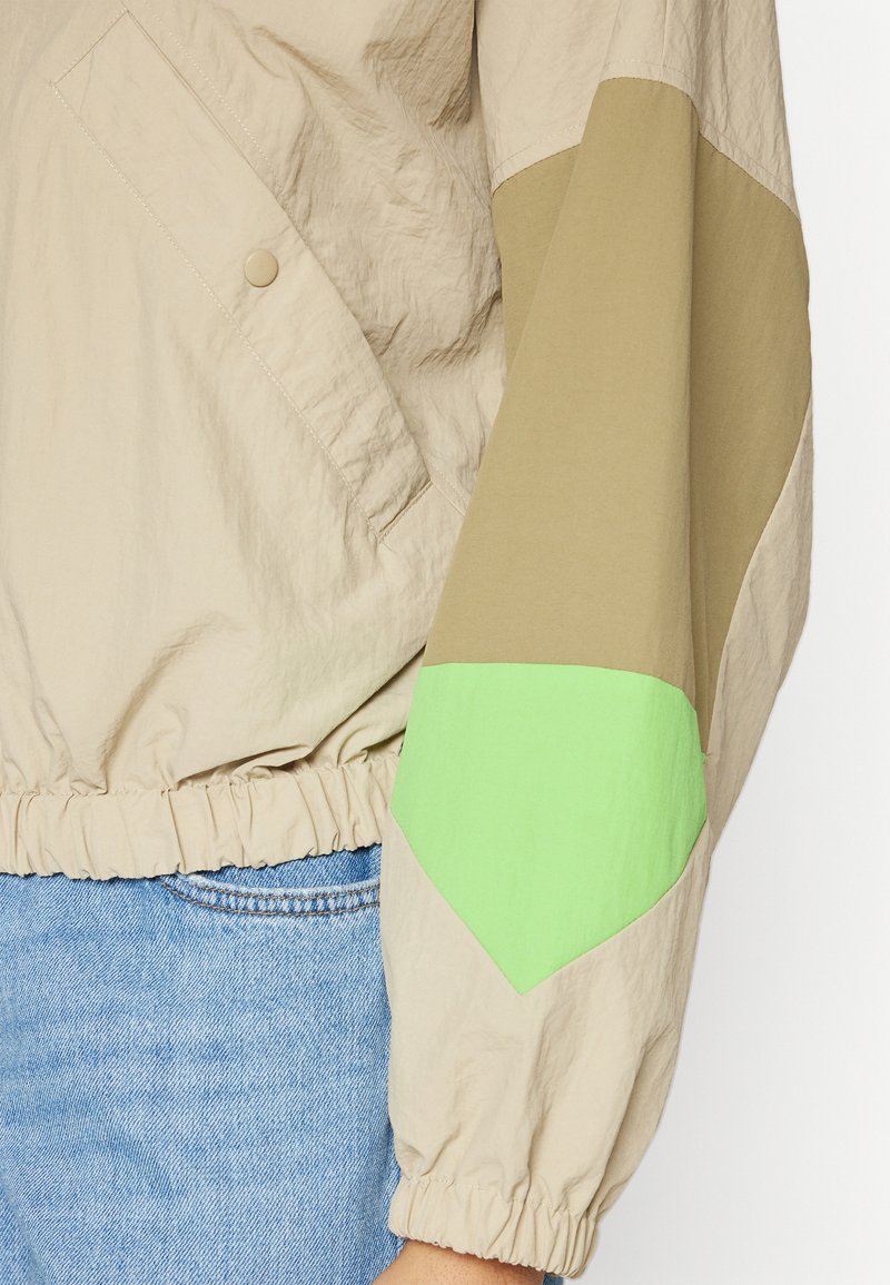 Chaqueta beige con dobladillo elástico, que presenta un panel en los hombros de color marrón contrastante y verde brillante. El material es ligero y tiene un acabado texturizado.