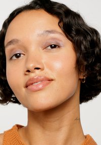 Primo piano di una persona con capelli corti e ricci e pelle beige. Hanno ombretto viola luccicante, labbra glossy e un orecchino a cerchio d'oro.