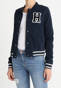 Námořnická modrá bomber bunda s bílými proužky na manžetách a spodním lemu, s bílým logem "H" na hrudi a bočními kapsami. Měkká textilní struktura.
