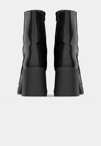 Černé kotníkové boty s elegantním, lesklým povrchem, ozdobené zipem na zadní straně a robustním, texturovaným podpatkem.