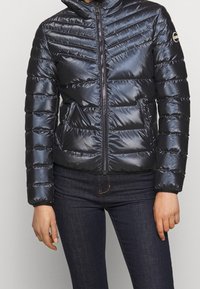 Laivaston sininen toppatakki, jossa siksak-mallinen tikkaus, korkea kaulus ja vetoketju edessä. Takissa on sileä, kiiltävä pinta ja sivutaskut.