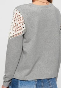 Graue Sweatshirt mit langen Ärmeln, mit aufwendigen weißen Häkel-Details an den Schultern. Weicher Stoff und lockere Passform.