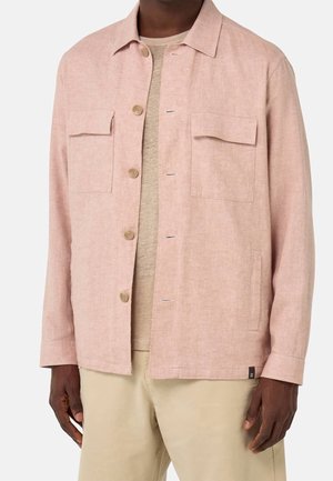 Uomo che indossa una giacca-camicia rosa chiaro con due tasche sul petto sopra una maglietta beige e pantaloni beige, in piedi davanti a uno sfondo bianco.