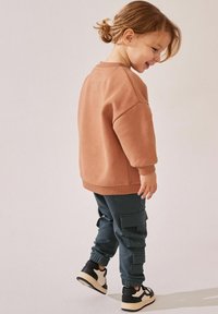 Sweter w jasnobrązowym kolorze z prążkowanymi mankietami, zestawiony z ciemnozielonymi spodniami cargo z bocznymi kieszeniami. Czarno-białe sneakersy dopełniają outfit.