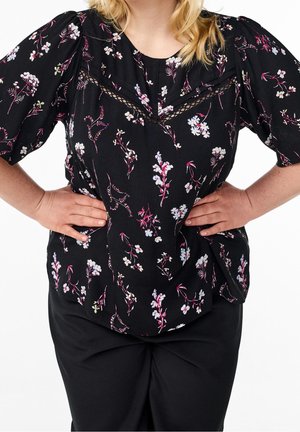 Kvinde iført en sort blomstret bluse med blondedetalje, stående med hænderne i siden, beskåret under hagen og over knæene.