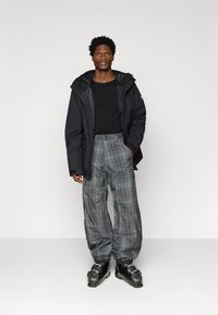 Moški v črni jakni, črni srajci, sivih kariranih snežnih hlačah in črnih smučarskih čevljih, stoji ob enobarvni svetli ozadju.