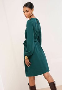 Smaragdovo zelené šaty z mäkkej tkaniny, s dlhými rukávmi, opaskom v páse a detailom na chrbte v tvare kľúčovej dierky. Hnedé čižmy po kolená.